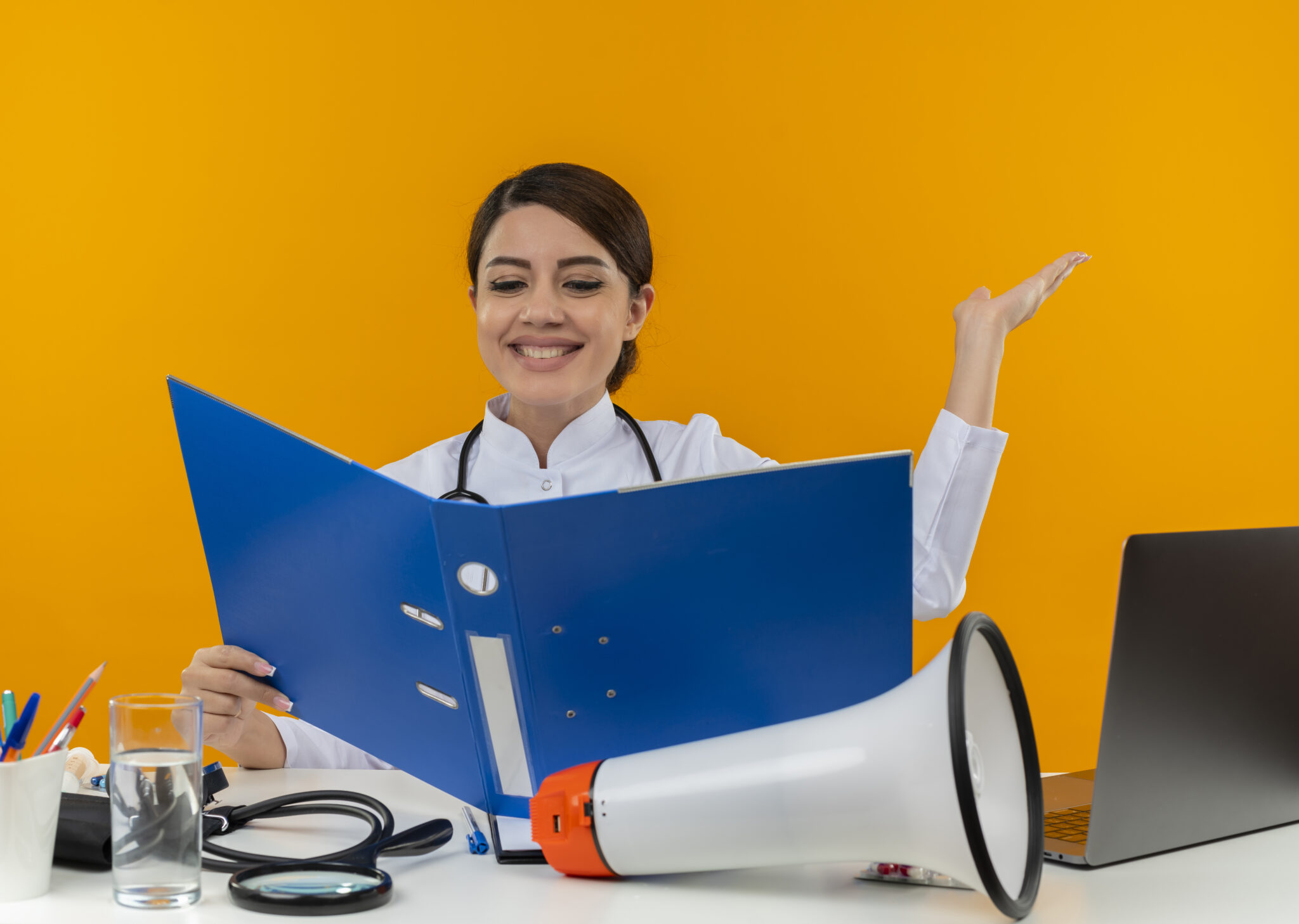 smiling young female doctor wearing medical robe with stethoscope sitting at desk work on computer with medical tools holding and looking at folder and points with hand to side on isolation yellow background